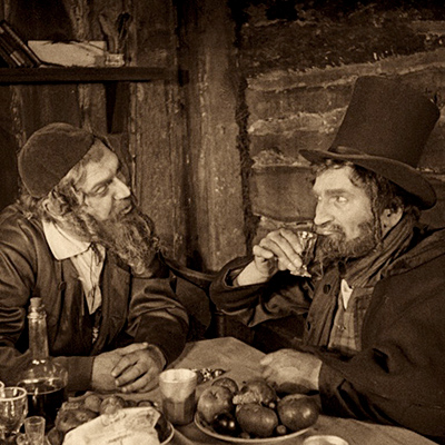 Sepia-toned still from The Ancient Law (1923). Two men with sidecurls seated at a table with food and drink, in conversation. One is wearing a kippah, speaking to the other who is wearing a top hat and taking a sip from a small cup.