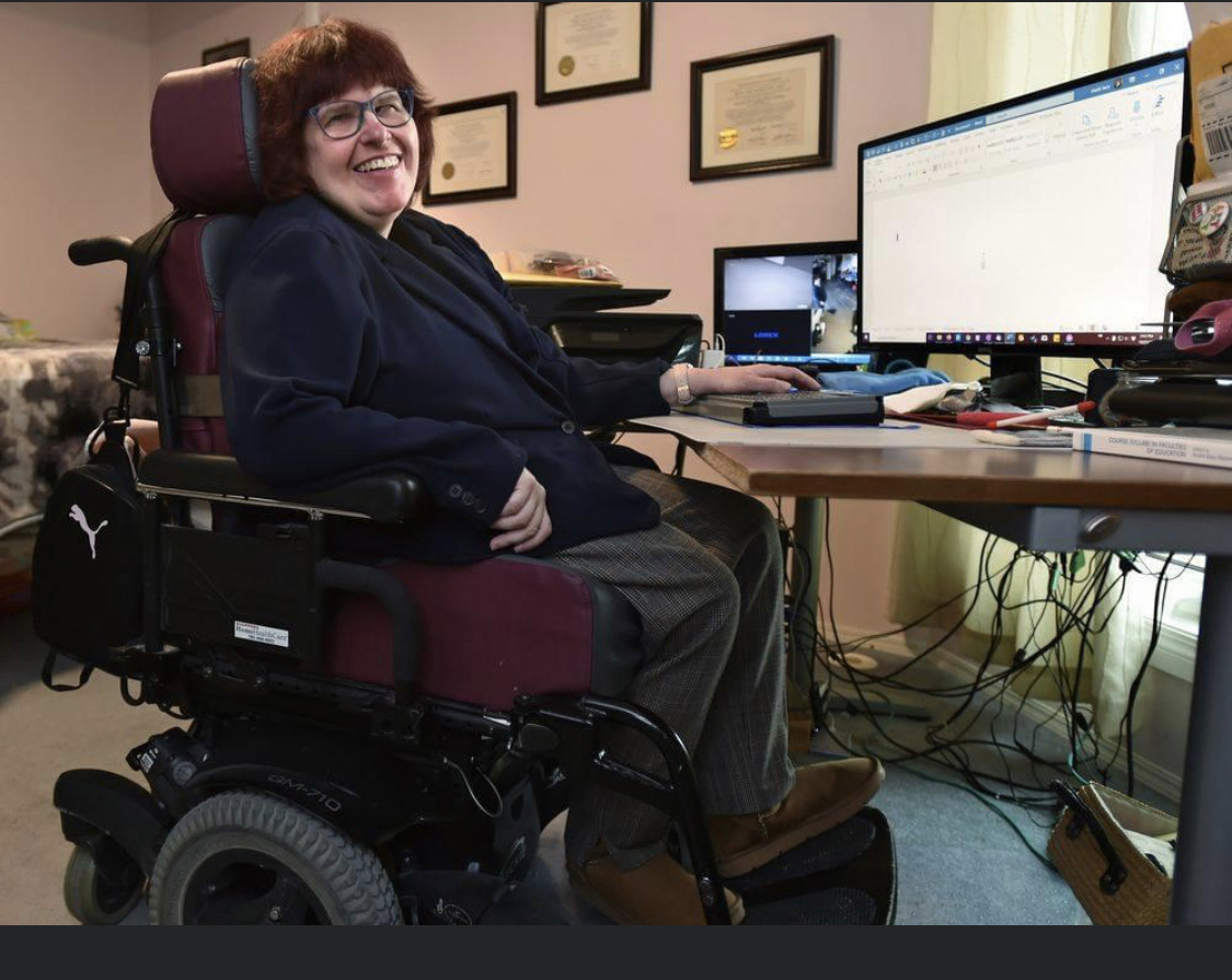 Heidi Janz wearing a navy blue blazer and glasses, at a computer table on a power chair, in a room with translucent curtains filtering the sunlight and framed gold-sealed certificates on the wall