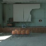 Still from Mariupolis (Mantas Kvedaravicius, 2016). Furniture and fixtures in an auditorium are in disarray. Debris litters the floor and stage. Sections of row seating are pushed aside. A single bed frame is under a torn projection screen hanging from the ceiling beside a de-installed television. Windows and a hole in the wall let sunlight in.