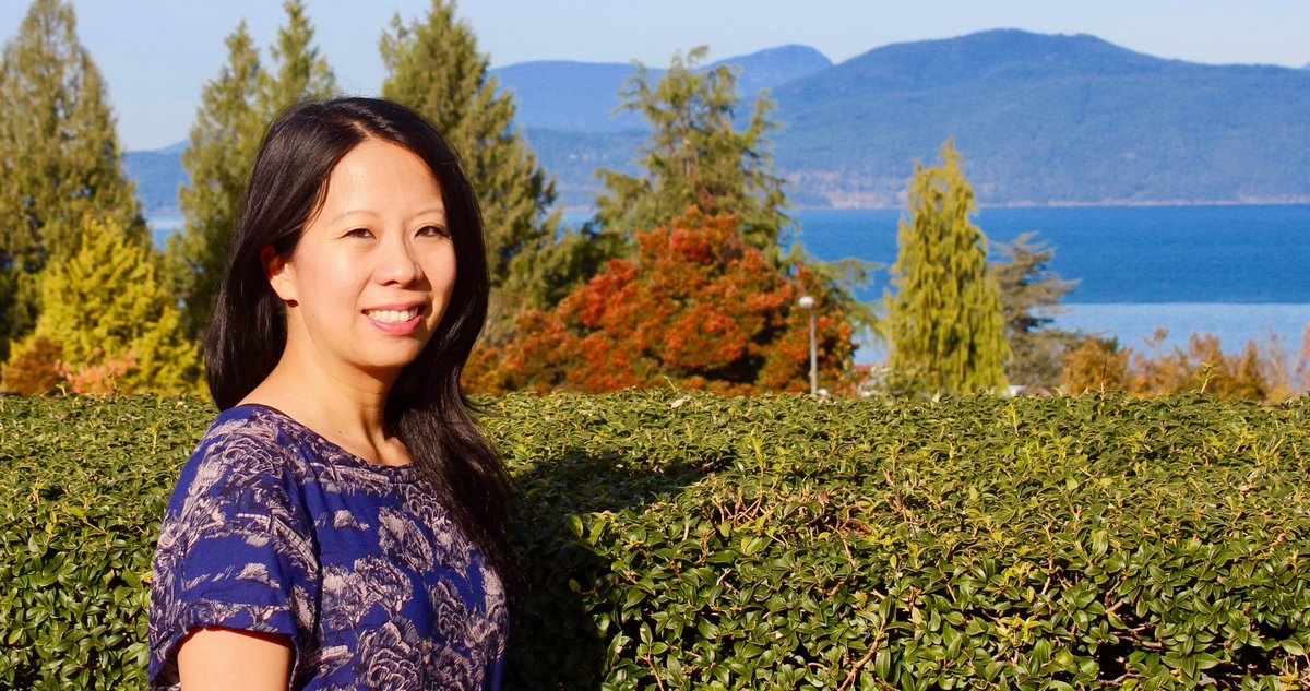 Y-Dang Troeung is smiling on a sunny day in the UBC Rose Garden with trees, water, mountains in the background