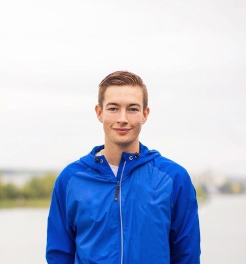 Dylan Annandale wearing a blue zip-up jacket, smiling, with low trees along a shoreline in the background