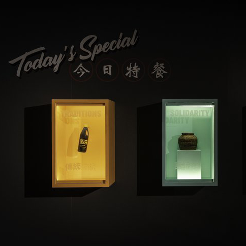 ​​Black gallery wall with “Today’s Special / 今日特餐” lettered above three display cases holding contemporary and older condiment and food vessels and words, translucent, on the plexiglass front, including “Traditions / 傳統” (in Traditional Chinese), and “Solidarity”. Detail from A Seat at the Table exhibition at the Museum of Vancouver.