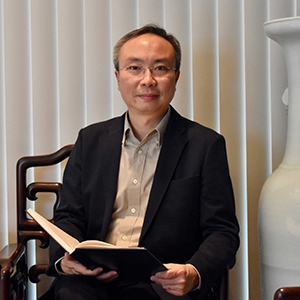 Professor Sze-Wing Tang wearing a taupe button-down collared shirt and black blazer and glasses, smiling, seated in front of a window shuttered with vertical blinds beside a large white vase, holding an open hard-bound book on his lap