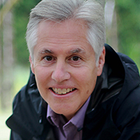 Professor John Borrows wearing a light purple collared shirt and black coat, smiling, in front of greenery