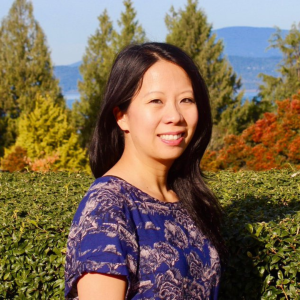 Y-Dang Troeung is smiling on a sunny day in the UBC Rose Garden with trees, water, mountains in the background