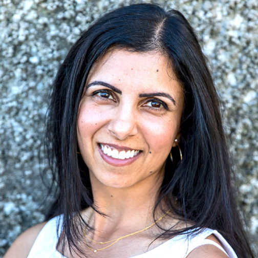 Renisa Mawani smiling, wearing a white top, with concrete wall in the background