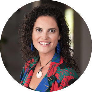 Tiara Na'puti is smiling into the camera. Tiara has brown curly hair, blue earrings, and a colorful blazer on